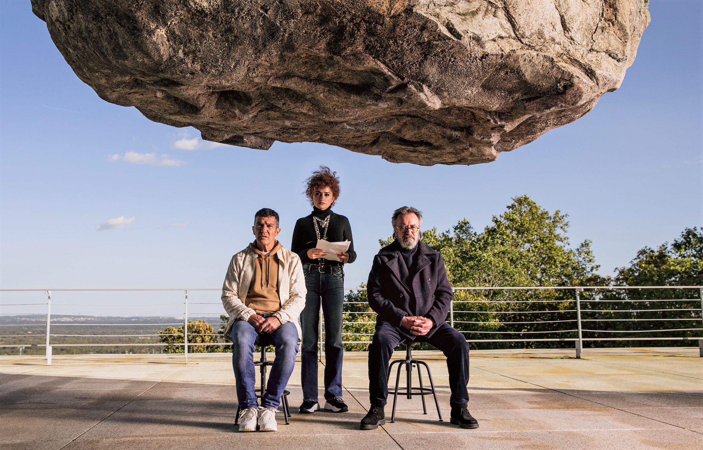 A woman stands and two men sit on chairs underneath a suspended big rock,