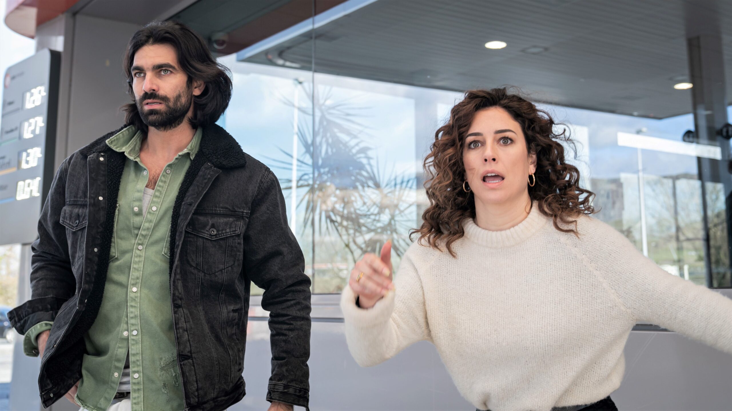 A bearded man and a dark-haired woman looking stressed and anxious outside a building